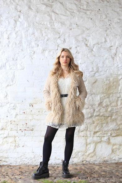 centred against a whitewashed masonry wall is Alice Louise, a model. She wears a hairy coat over a white belted shift over black tights and Doc Marten or similar boots. A blonde Goth.  The floor is cobbles.