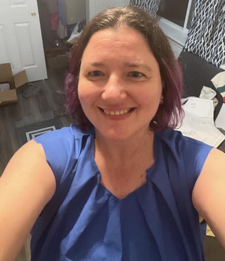Smiling woman wearing a sleeveless blue dress with a pleated neckline, selfie angle only shows face and upper bust