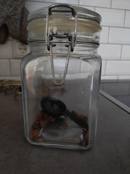 Photo of a glass jar, that contains a plastic measuring spoon and only a few, scattered and lonely looking coffee beans.