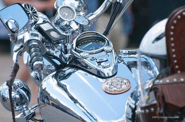 A closeup of the front of a motorcycle, looking at its area between the handles.  Lots of really shiny chrome, with mirrors and instruments.  A round medallion with an Indigenous art pattern has been placed between the speedometer and driver.  And ahead of the speedometer is mounted an old school iPod, in a black case with plastic over the round dial and the display area.