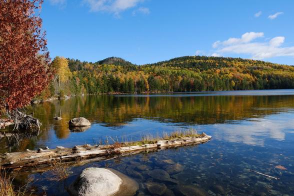 A sunny fall day, a lake with clear water, surrounded by forested hills in yellows and green. Blue sky with small clouds mirrored in the lake.
