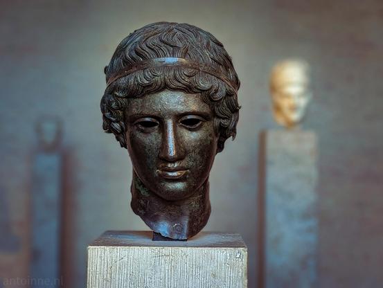 A prominent ancient Greek bronze sculpture of a youth's head, styled in the manner of the Severe or Early Classical period (circa 5th century BCE). The sculpture depicts a young, beardless male figure with finely articulated, wavy hair bound by a simple headband. His expression is calm, dignified, and idealized, characteristic of classical Greek bronze casting.

In the softly blurred background, other museum display pedestals and sculptural fragments are visible, indicating an exhibition setting within an antiquities museum.

They are displayed at the Glyptothek in Munich, Germany.