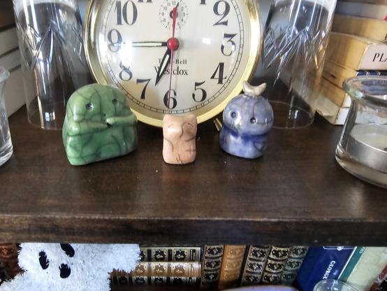 Three tiny raku ceramic spirits stand at attention in front of an old alarm clock. 

First a stout green one with a pensive look on its friendly little face, then a little pink fox figure, and finally a blue figure with a golden crescent moon on its head. They're raku ceramic, with the glaze spiderwebbed with a pattern of fine sooty cracks. 

From the shelf below, a festive ghost peeks into the frame.