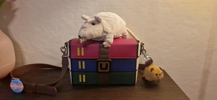 a winrar bag with a rat plushie on top, a trans pin on the strap and a… hamster plushie? next to the other side of the strap