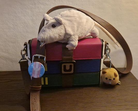 a winrar bag with a rat plushie on top, a trans pin on the strap and a… hamster plushie? next to the other side of the strap