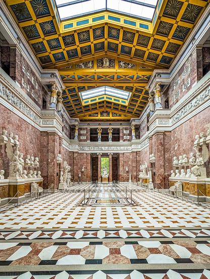The grand Neoclassical interior of the Walhalla memorial, a hall of fame located above the Danube River in Donaustauf, near Regensburg, Bavaria, Germany. 

Built by King Ludwig I of Bavaria to honor notable figures of Germanic history, culture, and science, the hall is richly adorned with reddish marble walls, decorative friezes, and a striking coffered ceiling accented in blue and gold with star motifs, illuminated by glass skylights.

Rows of white marble busts representing prominent historical figures line the lower walls on pedestals and shelves. Sculpted figures, winged victories, and caryatids accent the upper galleries and structural tiers.
