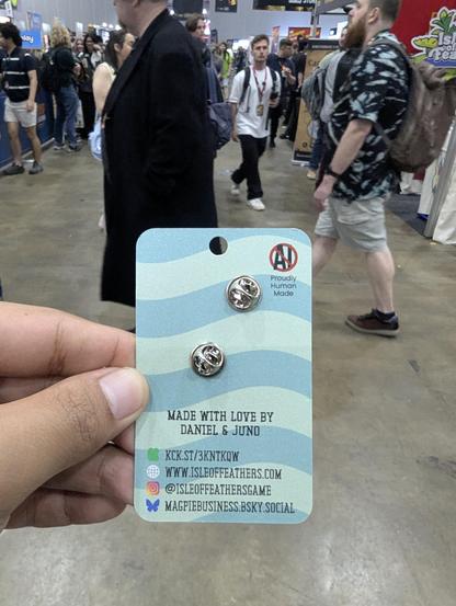 A left hand holding the backside of the enamel pin, showing the name of the makers and that they didnt use AI on it.
