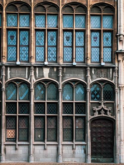 This detailed historic building facade features classic Northern European Gothic/Renaissance architecture, characteristic of historic guildhouses found at the Grote Markt in Antwerp.

The upper and lower levels feature large, multi-pane mullioned windows with intricate diamond-shaped leaded glass panes. The upper-row windows showcase delicate painted coats of arms or medallions embedded within the glass.

On the lower right, a heavy dark-brown wooden door features rows of circular decorative studs and brass hardware, framed by an ornate, flamboyant stone archway and sculptural details above.