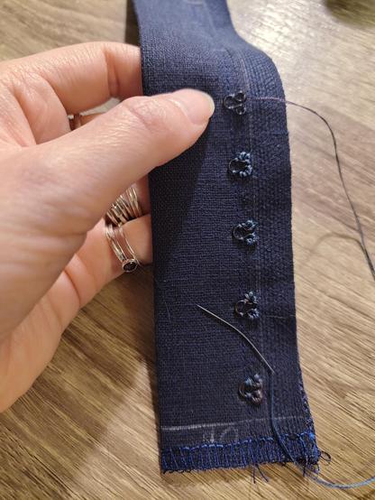 Close up photo of dinky little eyes of the hooks and eyes variety being sewn onto a strip of doubled over dark blue linen. There's a line of straight stitch onto which the visible, needed for use part of the eyes are placed. The handles are sewn on with a lighter blue thread and a tiny curved sewing needle. The topmost eye has only been stitched in place in four connection points, to keep it still while the handles are addressed.