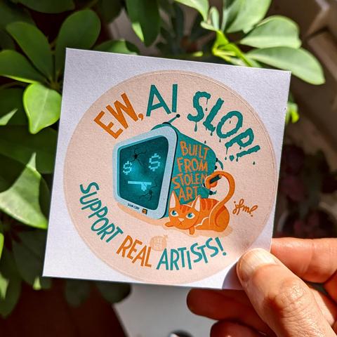 Photo of a hand in front of a green plant holding a sticker on a square piece of paper showing an illustration in ochre and mint green of a tv-like robot's head that has dollar signs for the eyes and two bullet holes in it's display, making a :P emoji. Laying at it's side is a yellow cat, looking angry that is grabbing a marker with her tail. She's just written „Built from stolen art“ on the side of the robot . Above the illustration it says „Ew, AI Slop!“ and beneath it „Support Real Artists!“, with a little speech bubble pointing at the cat, saying „like her“.