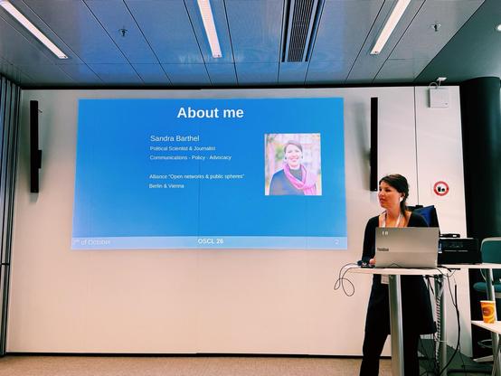 a photo showing Sandra Barthel (samvie) behind a podium, speaking, while there is a slide on a big screen behind her with her photo and her "about me" information