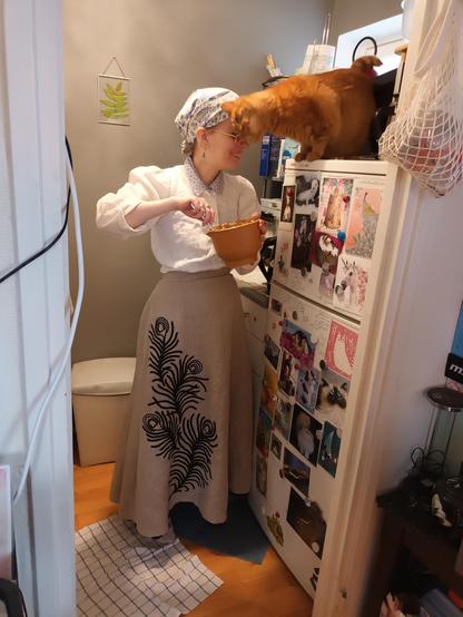 A very sweet photo, through a doorway, of Sini standing in her very small kitchen mixing pastry filling in a small plastic bowl. On top of a fridge, in front of a microwave, is the blurry shape of a ginger tabby cat headbutting her in the forehead, his whiskers to the front and with an expression of bliss on his little kitty face. She is grinning, eyes downcast. Both are warmly lit from above. The fridge is covered in colourful postcards and she's wearing a white linen blouse with the sleeves rolled up, a blue and white scarf on her head in the style of a laundress, and a nice tan linen skirt with elaborate peacock feather soutache in black. It's all very eclectic and cheerful!