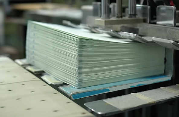 A tall stack of thread-sewn notebook pages rests under a mechanical press along a factory assembly line.