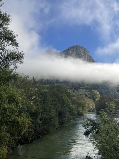 The image shows a river flowing through green vegetation following the direction of a mountain that is partly hidden by the morning haze. Behind the haze the sky is blue