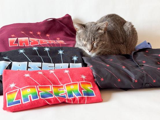 A grey tabby cat snoozes surrounded by a pile of laser-themed shirts