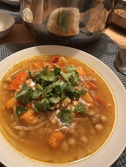 A plate of clear soup with chunks of roasted pumpkin, chickpeas and strips of lemon peel. Garnished with yoghurt, crispy chickpeas and fresh coriander. Plate sits on a placemat with a spoon next to it. Reflect can be seen in a big soup pot.