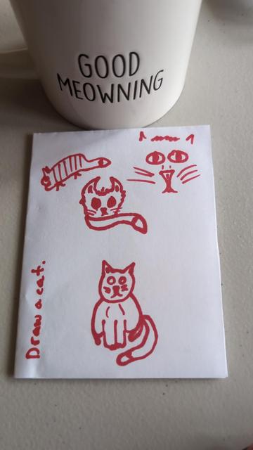 A folded piece of printer paper sits below a "Good Meowning" mug of tea. On the paper are several perfectly drawn cats in pink marker, along with the words "Draw a cat".