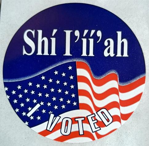 A color photo of a round sticker with the image of a US flag waving in the bottom half. On a blue background in white lettering on along the top it says, "Shí I'íí' ah" and on the bottom it says, "I voted".