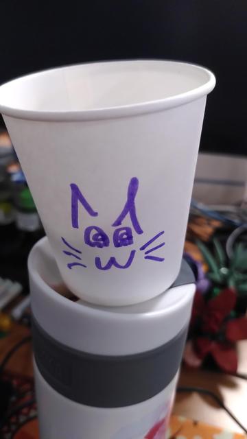 Photo of a papercup standing on top of a white cylinder with a gray grip ring. On the cup is crudely drawn a cat face made of 3 whisker on each side, a 3 for a mouth, two ^ for weara and two amongus for eyes (it's a largish arch with the opening on the bottom, an half circle flush with the bottom on the arch and also open on the bottom and another half-circle but roughly 2/3 high flush with the arch right side)