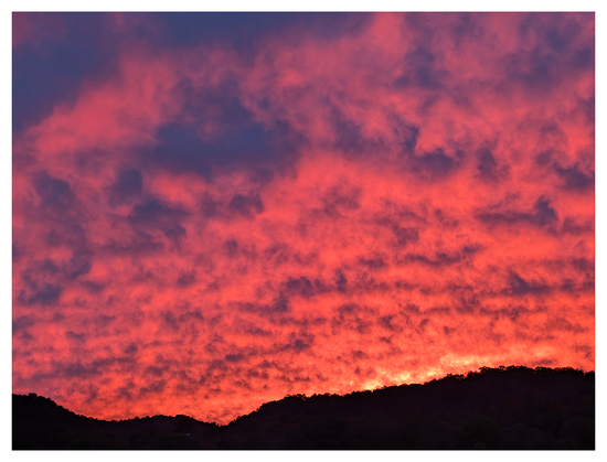 A vibrant sunset fills the sky with intense shades of fiery red, orange, and deep purple clouds. The dark, silhouetted shapes of rolling hills are visible along the bottom edge of the frame.