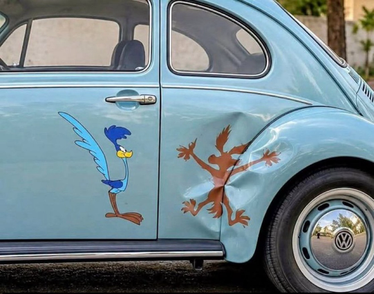 The The back quarter of a baby blue VW bug has a smashed back panel by the tire well. A cartoon image of a slatted Wile. E. Coyote adorns the crushed area, with a smiling cartoon image of the Road Runner on the door.