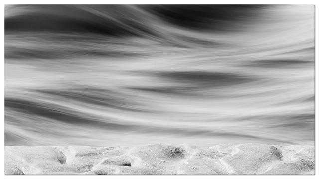 A black-and-white image showcasing smooth, flowing water above a textured sandy shore. The contrast highlights the movement of the water against the stillness of the sand.
