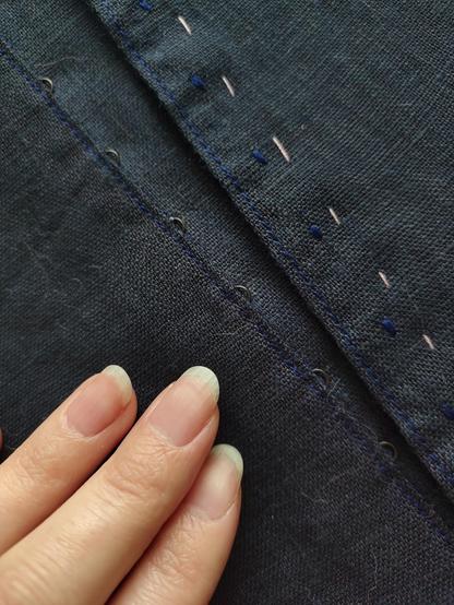 A close up photo of the side front placket and opening for a dark blue linen skirt. There is a tiny line of tidy topstitching in a slightly different dark blue thread right on the edge of both the eyes side, with the loop of the eyes peeking out only, and on the overlapping side, where the shadows of the body of the hooks is only slightly visible. There's a tidy bar tack at right where the hooks are attached, corresponding with the eyes on the other side: Otherwise the hooks are hidden. There's some basting thread along the placket, Sini's fingertips.