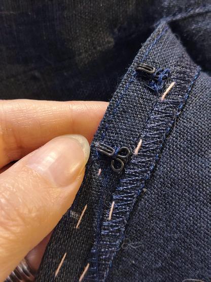 Very close up photo showing the sewing on of the hooks. The hook is set three millimetres from the edge of the opening, right at the line of the topstitching for even positioning. The one being sewn on is not yet fully covered, the one above it has been whipstitched all over, and has the bar tack through the handles, affixing it firmly in place through all layers.