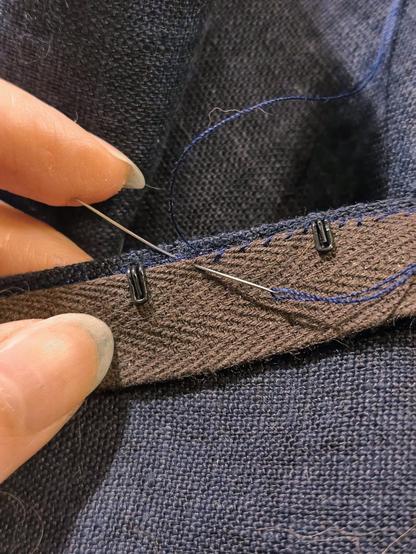 A brown herringbone cotton tape has been slid under the hook part of the hooks and the attachment part of them hiding absolutely all of the messy stitching, the seam allowance and other traces of construction. This is then being minutely whipstitched in place by hand, with dark blue thread. The sewing needle is incredibly fine, short and curved by use. The stitches are very very small and invisible from the right side of the garment.