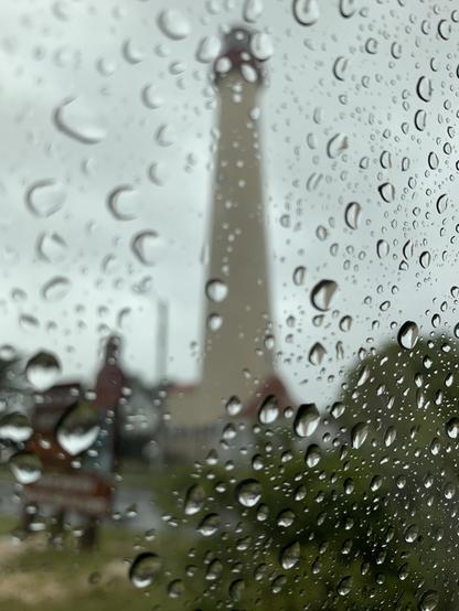 A photo taken in 2019 of the lighthouse in Cape May, NJ, through a car window on a rainy day. Lighthouse is blurred, focus is on some of the drops of rain on the window.