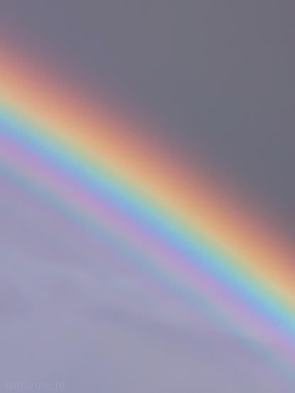 A section of a bright, vibrant rainbow extending diagonally across a stormy grey sky. A bold spectral arc featuring distinct color bands red, orange, yellow, green, blue, and purple, running diagonally from the upper left toward the lower right corner. Beneath the primary arc, faint additional repeating color bands are visible.