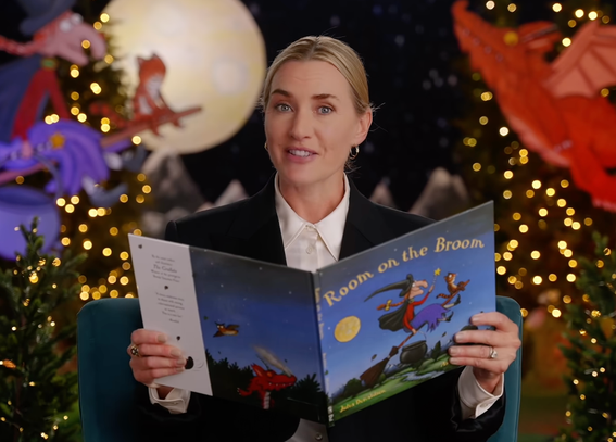 Actor Kate Winslet sits in front of a holiday backdrop holding open Julia Donaldson's illustrated picture book Room on the Broom.