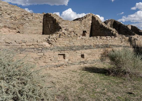A color landscape photo of ancient sandstone brick wall with four large rooms. There is no roof. The near wall is about half as high as the back wall. It extends across the center of the photo. There are four perpendicular walls extending from the shorter front wall to the taller back wall defining rooms. Two scrubby plants are on the right and left in the foreground. The sky is bright blue with puffy white clouds.
