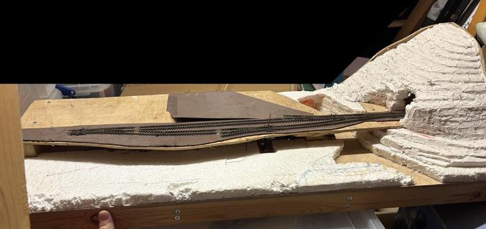 A model train layout featuring a section of track on a wooden base with white foam scenery. The layout includes a hill made of polystyrene covered in filler.