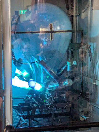 big glass orb, kind of lightbulb shaped, but massive, and glowing greenish blueish, sitting on a nest of brighter glowing glass tubes. A lot of human faces can be seen in the bulb!