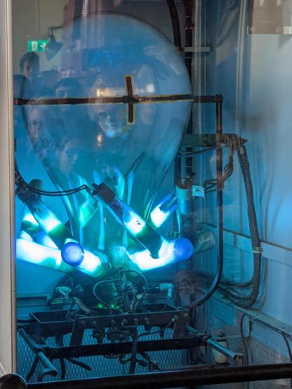 big glass orb, kind of lightbulb shaped, but massive, and glowing greenish blueish, sitting on a nest of brighter glowing glass tubes. A lot of human faces can be seen in the bulb!