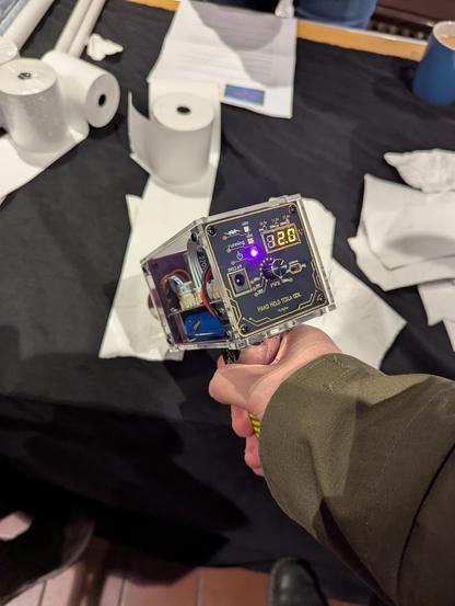 Outstretched arm pointing a handheld tesla coil towards a roll of paper. There's dials and sockets and LEDs on the back of the hand held Tesla coil