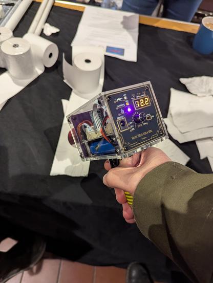 Another pic of an outstretched arm pointing a handheld tesla coil towards a roll of paper. There's dials and sockets and LEDs on the back of the hand held Tesla coil.  The tip of the coil can be seen here too.