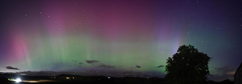 A wide night sky panorama showing an arc of bright Aurora rays in greens, purples and red hues