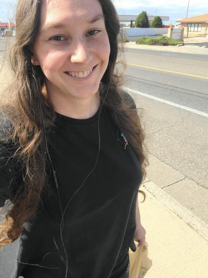 Me carrying a small grocery bag while walking on the sidewalk. I look tired, but I'm smiling and my hair has volume to it.