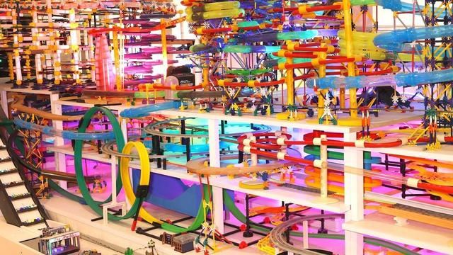 A massive, multi-tiered marble run constructed from colorful toy tracks, plastic scaffolding, and looping roller coaster sections spans across several white shelves.