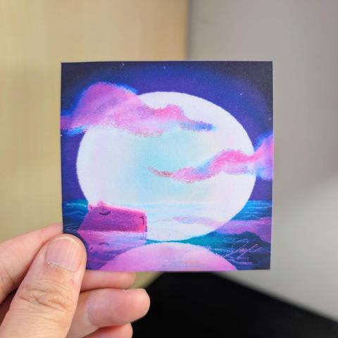 Photo of a left hand holding a square sticker. The sticker is a painting of a capybara resting in water with a full moon in the background.