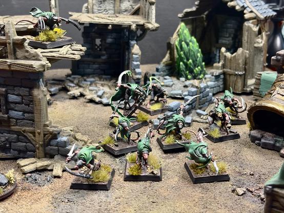 Painted cities of ash Skaven Gutter runners with green cloaks and long curved claws, arranged in a skirmish line on tufted-grass bases amongst a ruined city. Weathered ruins of stone-and-timber buildings surround them; one figure stands on an elevated wooden platform at upper left, a large chunk of warpstone emerges from a ruined building in the background. Dark neutral background, shallow depth of field.