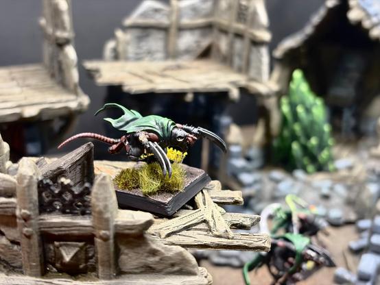Close-up of a Gutter Runner with a green cloak, pink striped tail and long scythe-like claws, standing on a base topped with a tuft of yellow-green grass. They’re perched on the balcony of a ruined building. The background is softly blurred, showing more ruins, a warpstone chunk more gutter runners below.
