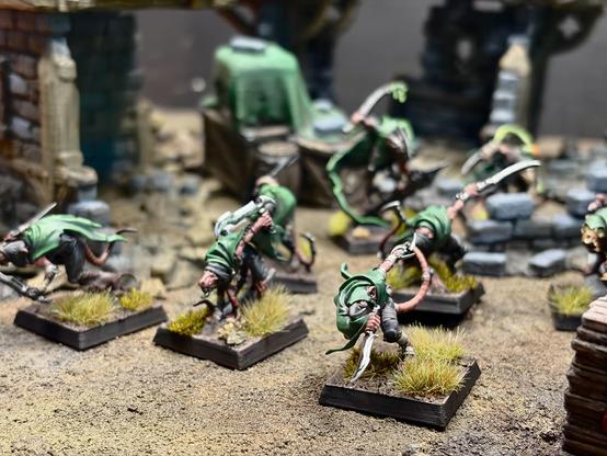 Eight painted Gutter Runners with green cloaks, pink tails and shining blades, spaced out on grass-tuft bases across a rubble-strewn sandy surface. A green-draped crate sits in the blurred background against stone ruins; a stack of wooden planks is visible at the lower right edge. Low-angle macro shot with shallow focus.