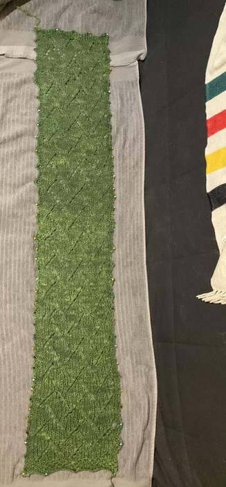 A green knitted scarf with a zigzag pattern is pinned flat to two grey towels to shape it