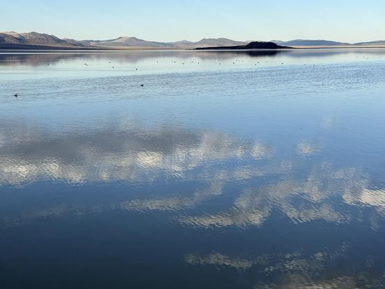 A serene lake surrounded by a mountain range. The water reflects the sky and clouds, like a frosted glass.