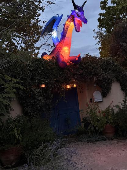 An inflatable dragon in vibrant colors is perched atop an adobe entry gate, surrounded by greenery. The dragon has blue wings and emits a warm glow, contrasting with the blue doors below it. The setting is dusk, adding a magical atmosphere.