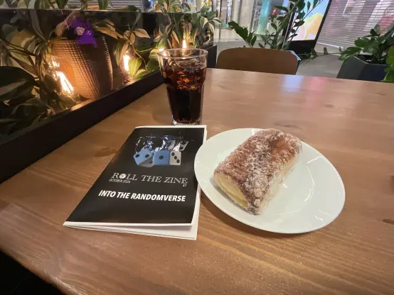 A copy of Roll the Zine 7: Into the randomverse on a cafe table next to a glass of coke and a sugary pastry.
