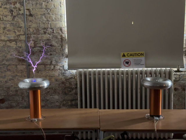 Two smaller Tesla coils on a table. Each has a vertical rod throwing off forked sparks. They’re both playing a tune together. All the other ones playing tunes were individual coils, so this was quite neat.